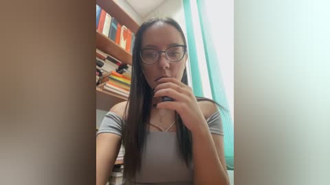 Media: Video of a young woman with long dark hair, wearing glasses, a gray off-shoulder top, and a contemplative expression, sitting indoors. Background features a bookshelf and teal curtains.