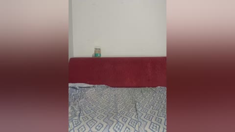 Video of a simple, empty room with a red cushioned headboard, blue and white patterned blanket, and a small glass jar on a white wall.