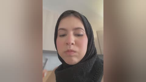 Media: Video of a woman with medium brown skin, wearing a black hijab, looking down in a neutral indoor setting.