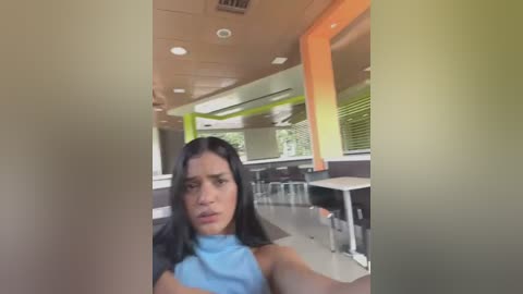 Media: Video of a woman with long black hair, wearing a light blue sleeveless top, sitting in a brightly lit fast-food restaurant with modern decor, green and orange accents, and a window view of greenery.