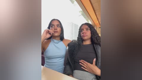 Media: Video of two young women with medium brown skin, one in a light blue sleeveless top and the other in a black sweater, standing side by side in a modern, bright interior.