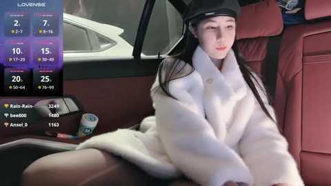 Media: Video of a young East Asian woman with long black hair, wearing a white fur coat, sitting in a red leather car seat. The background includes a car dashboard with various digital displays and a window showing a cityscape.