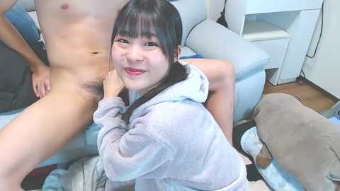 Media: A video of an Asian couple in a bedroom. The woman, with long black hair, is nude, wearing only a hoodie, and the man is partially visible, also nude. They are on a bed with plush toys.