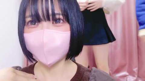 Media: Video of a young woman with short black hair, wearing a pink mask and a brown top. Another woman in a black skirt is partially visible in the background.