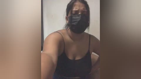 Media: Video of a woman with medium skin tone, wearing a black face mask, black spaghetti-strap top, and black hair tied back, indoors, blurred background.