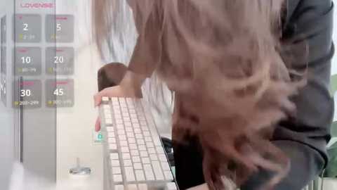 A video shows a woman with long blonde hair typing on a laptop in a modern office. The background features a calendar with highlighted dates. The image is slightly blurred.