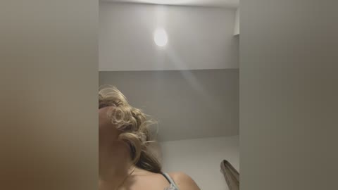 Media: Video of a blonde woman with curly hair, wearing a white tank top, lying on a bed with a bright ceiling light illuminating her face. The room is minimalist with beige walls and a simple design.