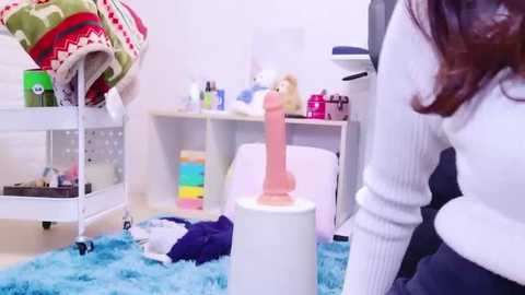 Media: Video of a young woman with long brown hair wearing a white sweater, standing near a white stool with a pink dildo and colorful toys scattered on a blue shag rug in a messy room.