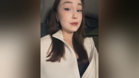 Video of a young woman with fair skin, brown hair in pigtails, and wearing a beige jacket over a black top. She has a neutral expression. Background is blurred, likely an indoor setting.