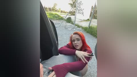 A video shows a young woman with bright orange hair and a red sweater lying on a gravel road beside a parked car. The background features a rural landscape with a sign and trees.