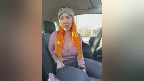 Video of a young woman with bright orange hair, wearing a pink cardigan and high-waisted pants, sitting in a car seat, looking surprised.