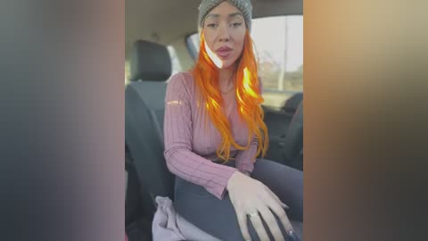 Media: Video of a young Asian woman with long orange hair and a gray beanie, wearing a pink sweater and gray pants, sitting in a car.