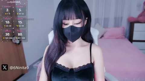 Media: A video of a young Asian woman with long black hair, wearing a black mask and lacy black top, in a bedroom with pink bedding.