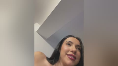 Video of a smiling, light-skinned woman with long black hair, possibly Latina, wearing pink lipstick, captured from a low angle, with a white ceiling and wall in the background.