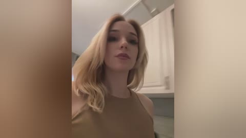 Media: Video of a young, light-skinned woman with long, wavy blonde hair and minimal makeup, wearing a beige tank top, standing in a bathroom with white cabinets and a towel.