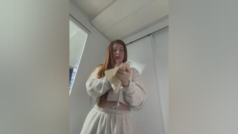 Media: A video of a young woman with long, straight, red hair, wearing a light gray hoodie and sweatpants, holding a tissue to her nose in a small, white room with sliding closet doors.