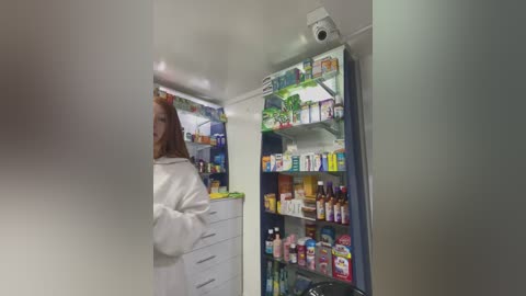 Media: A video of a white, middle-aged woman in a white lab coat, standing in a modern pharmacy with shelves filled with colorful medicine bottles and boxes.