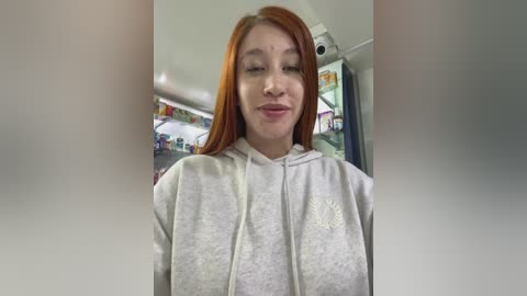 Video of a young woman with long, straight, auburn hair, wearing a light gray hoodie, smiling slightly. Background shows a cluttered room with colorful items and a ceiling fan.