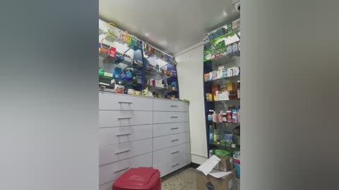 Media: Video of a cluttered, white kitchen cabinet with numerous medications, a red stool, and a speckled floor, capturing an unorganized, possibly medical storage area.