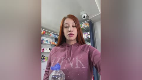 Video of a young woman with long red hair, wearing a pink hoodie, seated in a pharmacy. She holds a clear plastic water bottle. Background shows shelves of medications and a security camera.