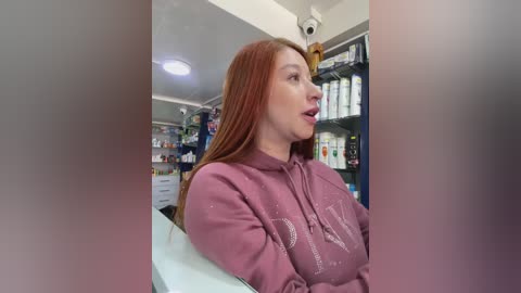 Media: Video of a fair-skinned redhead woman with long hair, wearing a pink hoodie, standing in a pharmacy. Shelves in the background are stocked with various products.