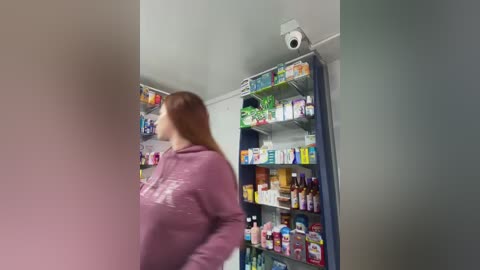 Media: Video of a woman with long red hair in a pink hoodie, standing in a well-lit pharmacy with shelves full of various products, including medicine bottles.