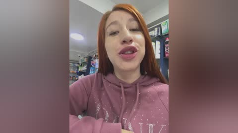 Video of a young woman with long, straight red hair, wearing a purple hoodie, winking and sticking out her tongue, in a brightly lit clothing store with shelves of clothes in the background.