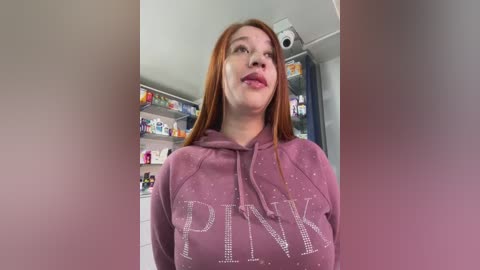 Video of a young woman with fair skin and long, straight red hair, wearing a pink hoodie with \"PINK\" in large, sparkly letters. Background shows a cluttered room with shelves filled with colorful items.