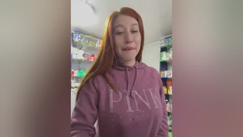 Video of a fair-skinned, red-haired woman in a purple \"PWN\" sweatshirt, standing in a well-lit pharmacy. Shelves in the background are stocked with various medications and products.