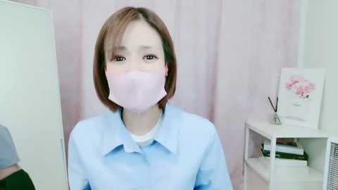 Media: Video of an Asian woman with short brown hair wearing a pink face mask and light blue shirt, standing in a pastel-colored room with a floral painting and white shelves.