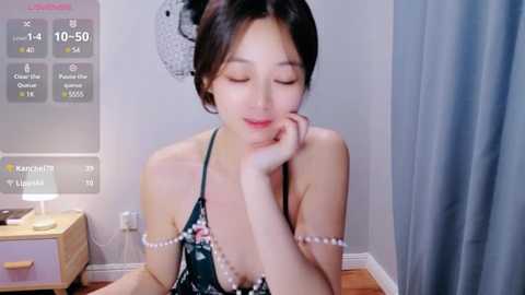 Media: Video of an Asian woman with fair skin and black hair in a floral dress, sitting at a desk in a bedroom. She's smiling and resting her chin on her hand.