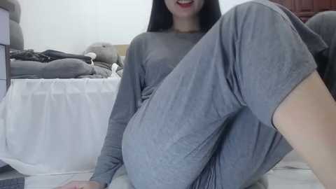 Media: Video of a smiling, dark-haired woman in a gray, long-sleeved shirt and pants, sitting on a white bed in a cozy bedroom with plush gray pillows and a wooden dresser.