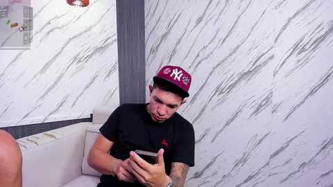 Media: Video of a young man in a black t-shirt and red New York Yankees cap, sitting on a white couch, holding a smartphone, in a room with white marble walls.