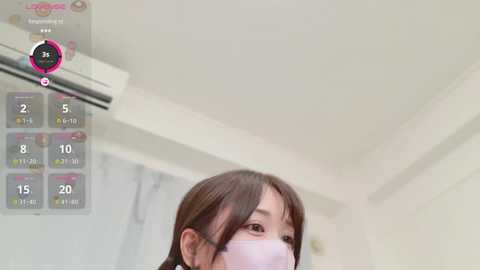 Media: Video of an Asian woman with shoulder-length brown hair, wearing a light pink face mask, standing indoors. Background shows a white ceiling and a partially visible air conditioner.