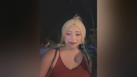 Media: Video of a smiling young Asian woman with fair skin, platinum blonde hair in a messy updo, wearing a red tank top. Background features a dark, blurred nighttime street with a few cars.