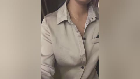 A video of a woman wearing a light beige, button-up shirt, partially unbuttoned, revealing a hint of cleavage. She has fair skin and is seated in a dimly lit room with a blurred background.