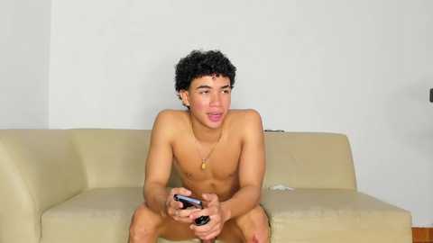 Media: Video of a shirtless, young, curly-haired Latino man with light brown skin, playing a video game on a beige couch against a plain white wall.