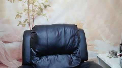 Media: A video of a black leather recliner chair with a cushioned headrest, set against a floral wallpaper featuring a green plant and soft pastel hues, with a white side table beside it.