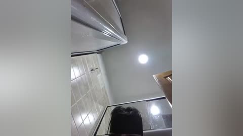 Media: Video of a modern, clean bathroom with white tiled walls, a glass shower enclosure, and a bright circular light fixture. A person with dark hair stands in the shower, partially visible.