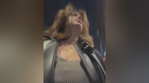 Media: Video of a blonde woman in a grey sweater and black leather jacket, slightly blurred, with a dark background, possibly an urban setting.