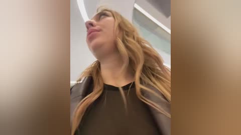 Media: A video of a young woman with long, wavy blonde hair, wearing a black leather jacket, gazing upward with a contemplative expression. The background features a modern, indoor setting with white walls and ceiling lights.