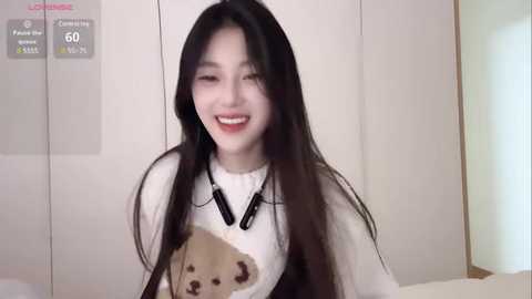 Media: Video of a smiling Asian woman with long black hair, wearing a white sweater with a teddy bear graphic, indoors.