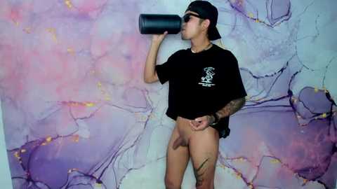 Video of a tattooed, shirtless man wearing a black cap and drinking from a black water bottle against a pastel marble-patterned wall.