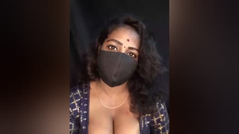 Media: Video of a dark-skinned woman with voluminous curly black hair, wearing a black face mask and a floral-patterned blue blouse with deep neckline, revealing ample cleavage, against a dark background.