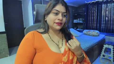 Media: A video of a South Asian woman with medium skin tone and long dark hair, wearing an orange blouse with a low neckline, in a modern bedroom with a bed, curtains, and furniture.