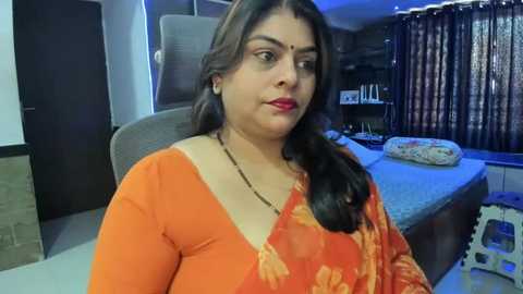 Media: Video of a middle-aged woman with dark skin and long black hair, wearing an orange floral sari, seated in a modern bedroom with a gray bed, dark curtains, and blue lighting.