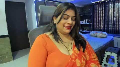 Media: Video of a middle-aged South Asian woman with long, straight black hair, wearing an orange floral sari, smiling in a modern, brightly lit room with a bed and dark curtains.