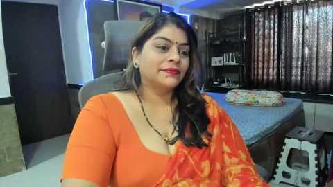 Video of a middle-aged woman with medium brown skin, wearing an orange sari with gold patterns, sitting on a grey chair in a modern bedroom with a bed, curtains, and a white chair in the background.