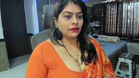 Media: Video of a South Asian woman with medium skin tone, black hair, and red bindi, wearing an orange saree, seated in a modern, cluttered bedroom with a bed, shelves, and curtains.