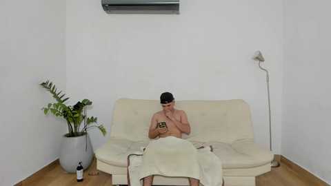 Media: A video of a shirtless man with dark hair, sitting on a beige couch, wrapped in a towel, holding a smartphone, in a minimalist room with a potted plant, air conditioning, and a floor lamp.